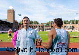 Kurtis Marschallas / Vida Press nuotr. - 4Active - 4Active Two male athletes shake hands on a running track at a stadium, one in a light blue jacket and the other in a teal sleeveless shirt with sponsor logos, with stands in the background.