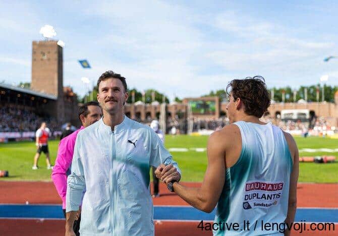 Kurtis Marschallas / Vida Press nuotr. - 4Active Two male athletes shake hands on a running track at a stadium, one in a light blue jacket and the other in a teal sleeveless shirt with sponsor logos, with stands in the background.
