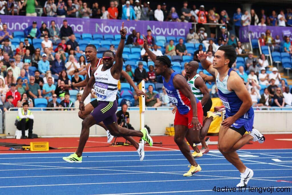 Four male sprinters race on a blue track with a cheering crowd in the stands.