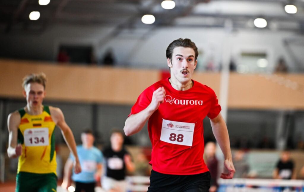 Male sprinter in a red shirt with bib 88 leading on an indoor track; opponent in yellow-green behind him.