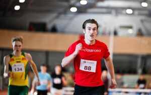 Male sprinter in a red shirt with bib 88 leading on an indoor track; opponent in yellow-green behind him.