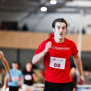 Male sprinter in a red shirt with bib 88 leading on an indoor track; opponent in yellow-green behind him.