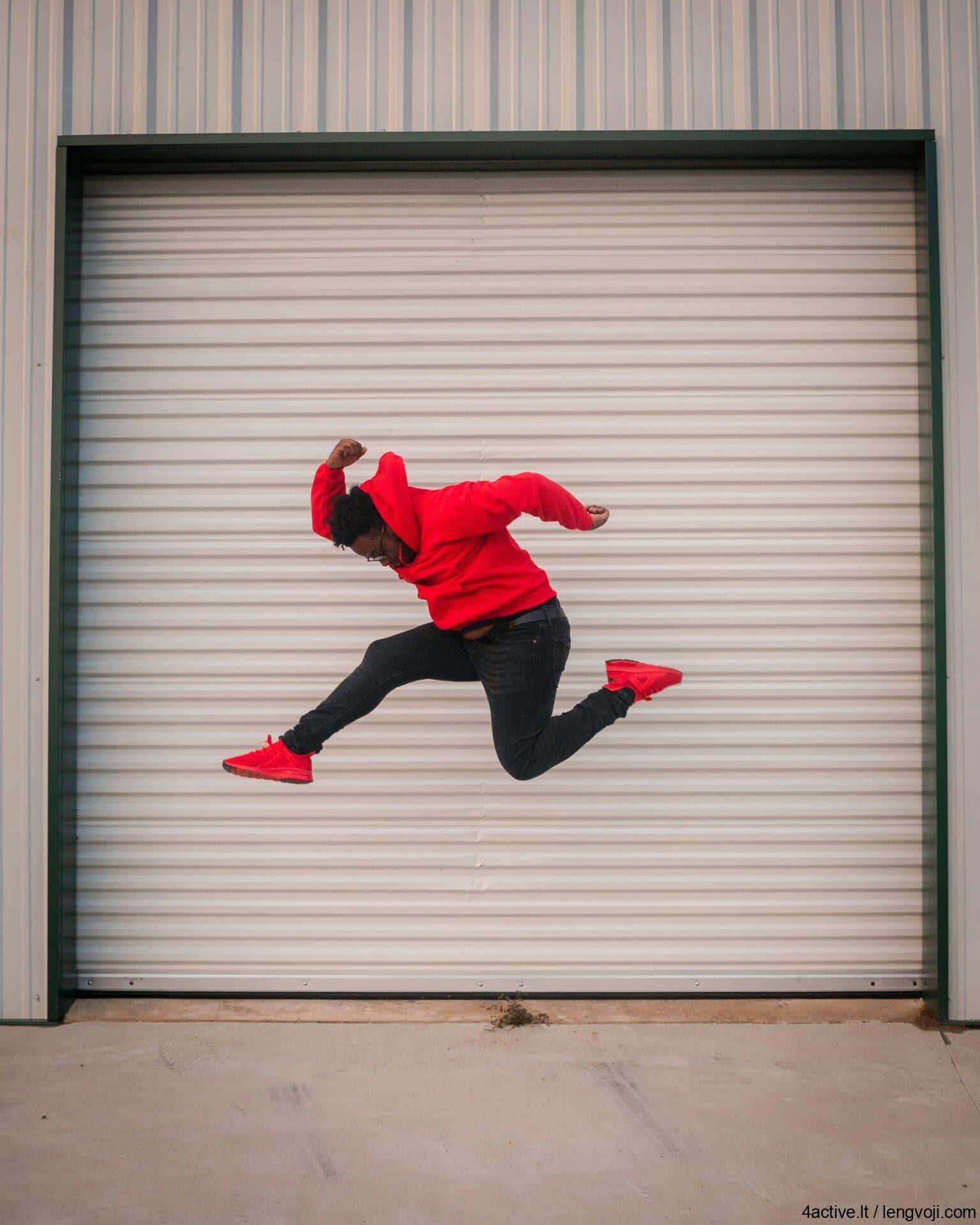 Person in a red hoodie and red sneakers jumps mid-air in front of a large white roll-up garage door.