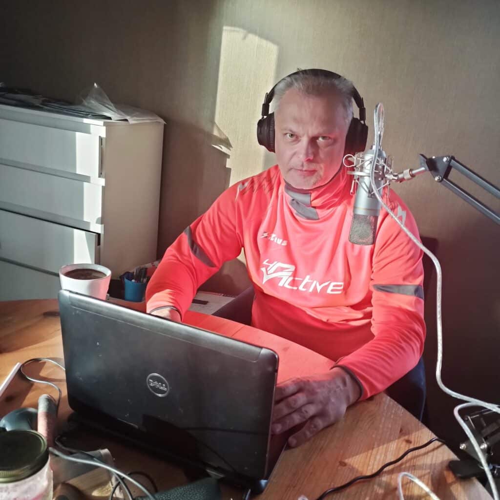 Man wearing pink athletic shirt with headphones speaks into a condenser mic at a desk setup, laptop open in front of him, coffee mug nearby.