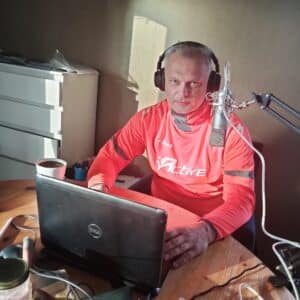 Man wearing pink athletic shirt with headphones speaks into a condenser mic at a desk setup, laptop open in front of him, coffee mug nearby.