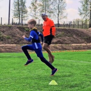 Young boy in a blue outfit sprints past a yellow cone on a grassy field, coached by a man in an orange jacket.