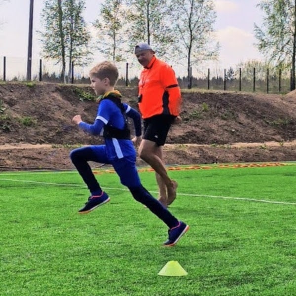 vilmantas ausra - 4Active Young boy in a blue outfit sprints past a yellow cone on a grassy field, coached by a man in an orange jacket.