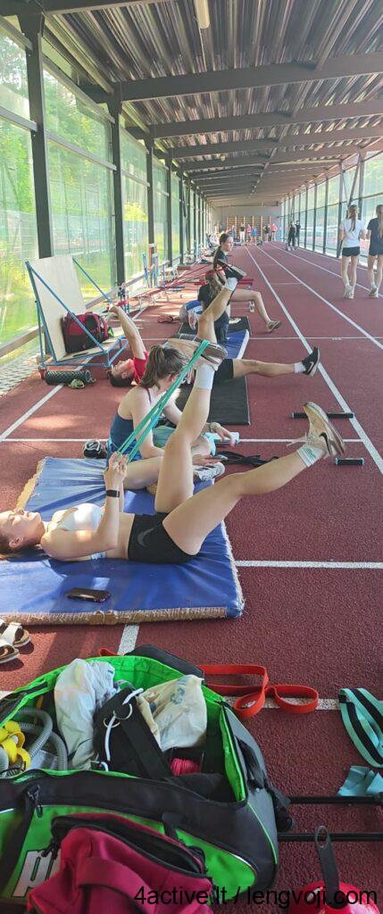 Indoor running track with athletes lying on mats doing leg stretches with resistance bands.