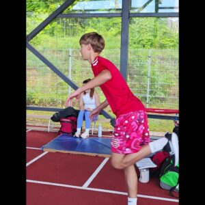 Boy in a red shirt and pink shorts running on an indoor track, with a girl seated on a bench in the background.