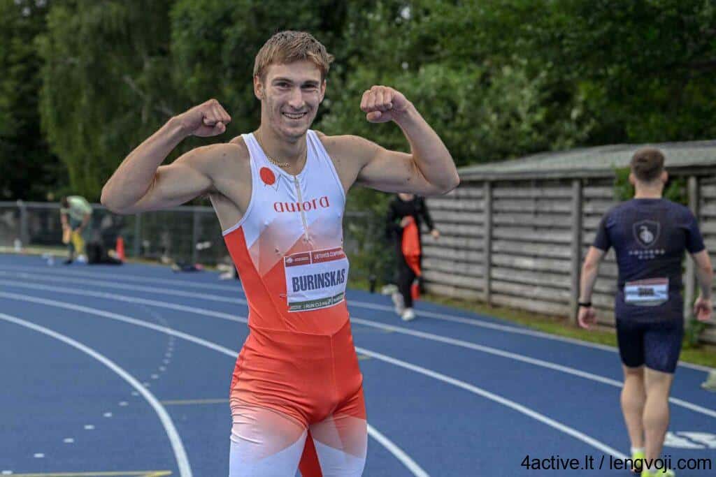 Sprinter on a blue track posing with flexed arms in victory, wearing a red‑white racing suit and bib 'BURINSKAS'.