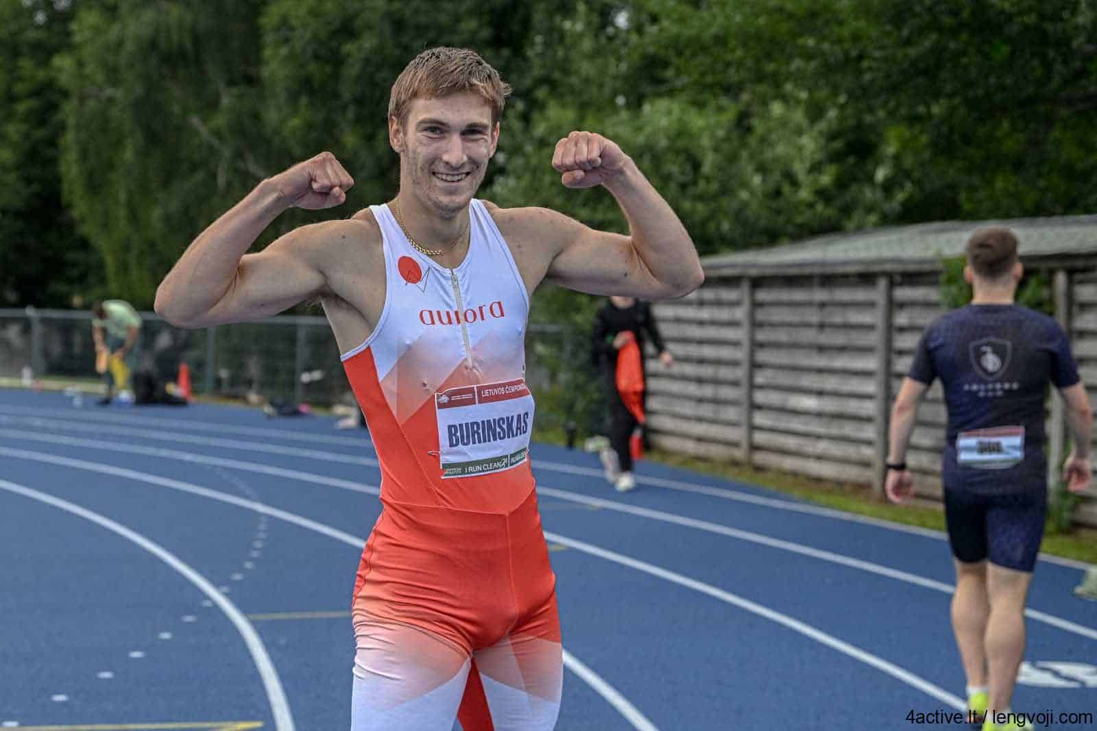 Sprinter on a blue track posing with flexed arms in victory, wearing a red‑white racing suit and bib 'BURINSKAS'.