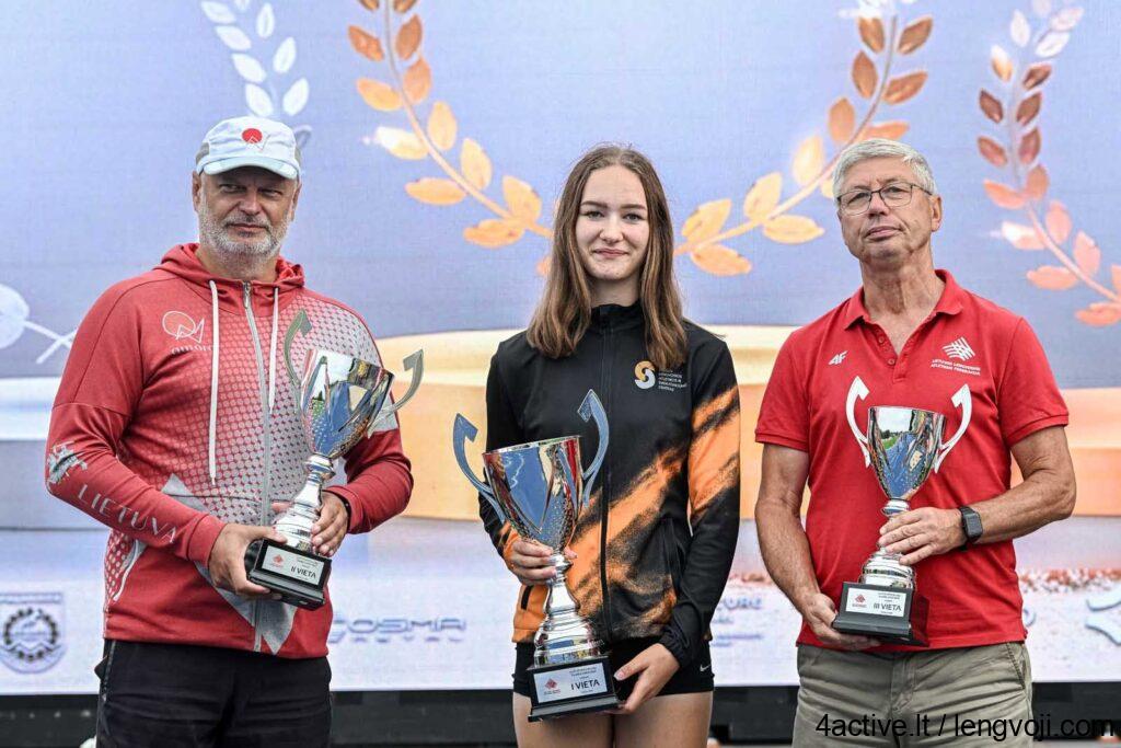 Aurora klubas - 4Active Three athletes stand on a podium holding large trophies, with a blue backdrop and laurel decorations behind them.