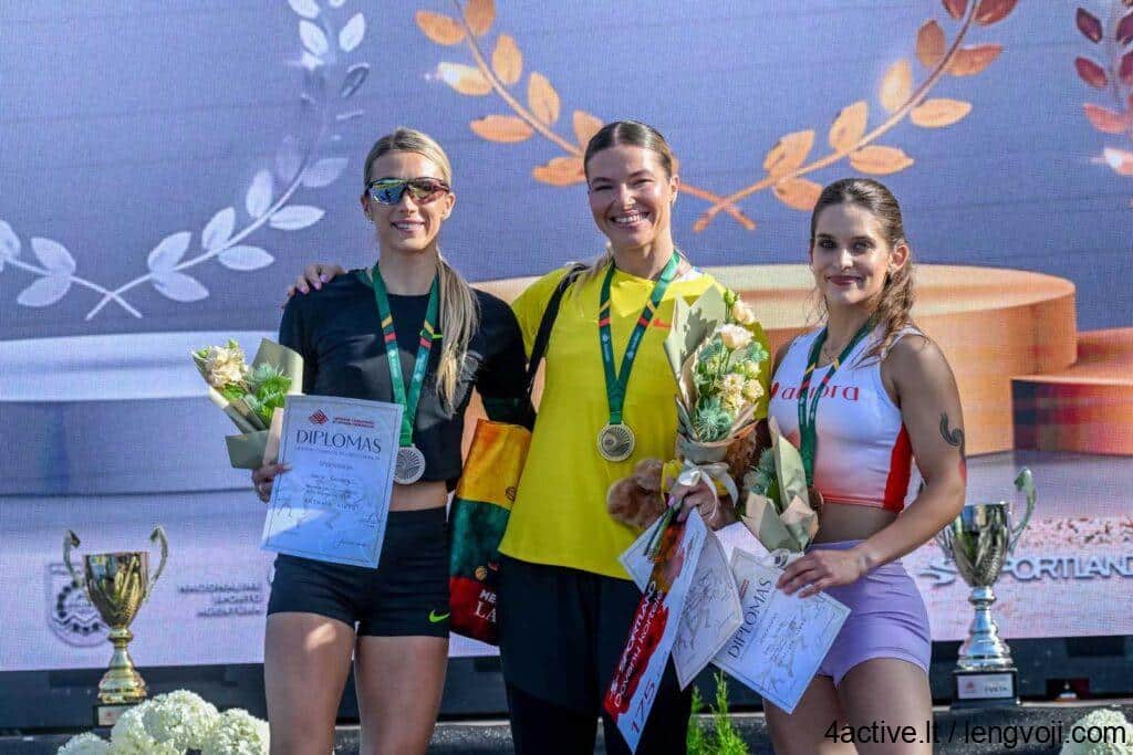 Diana_Veličkaitė_bronza_400m - 4Active Three female athletes on a podium wearing medals, holding bouquets and diplomas while smiling at the camera.