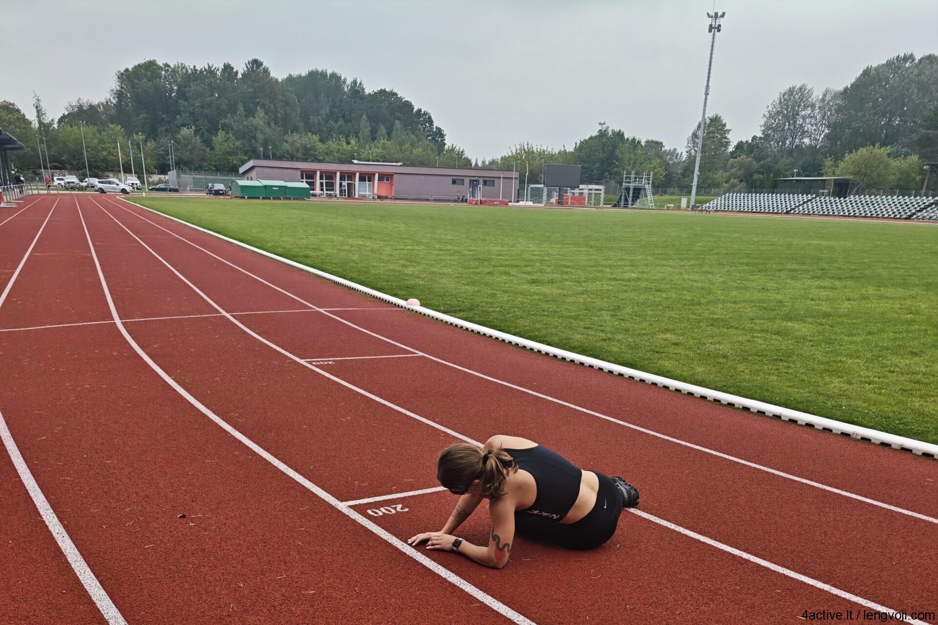 Athlete performing a push-up on a red running track beside a green field at a stadium.