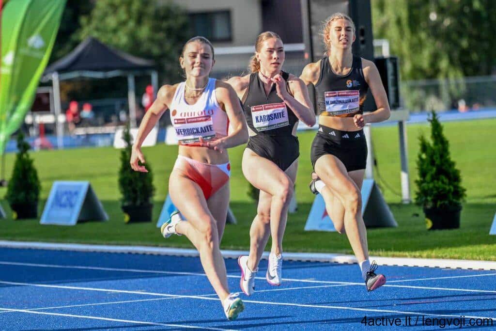 ieva_verbickaitė - 4Active Three female sprinters race side by side on a blue track outdoors, wearing bib numbers and athletic outfits.