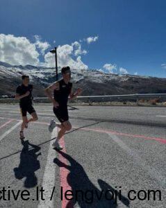 Two runners in black sprint along a sunny mountain road with a guardrail and snow-capped peaks in the background.
