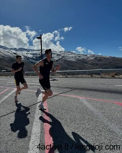 Two runners in black sprint along a sunny mountain road with a guardrail and snow-capped peaks in the background.