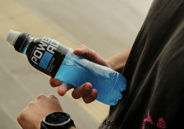 Energetiniai gerimai - 4Active A person holding a blue Powerade sports drink bottle outdoors, with a smartwatch visible on their wrist.